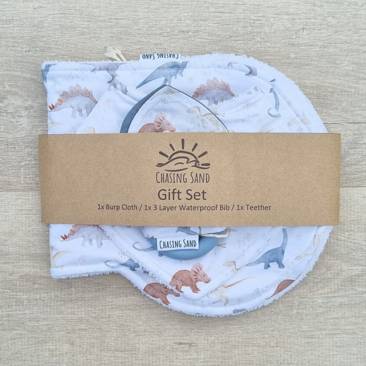 3 Piece Gift Set - Dino against wooden backdrop. Watercolour dinosaur illustrations white background. Each set contains 1x Burp Cloth, 1x Dribble Bib and 1x Teether.