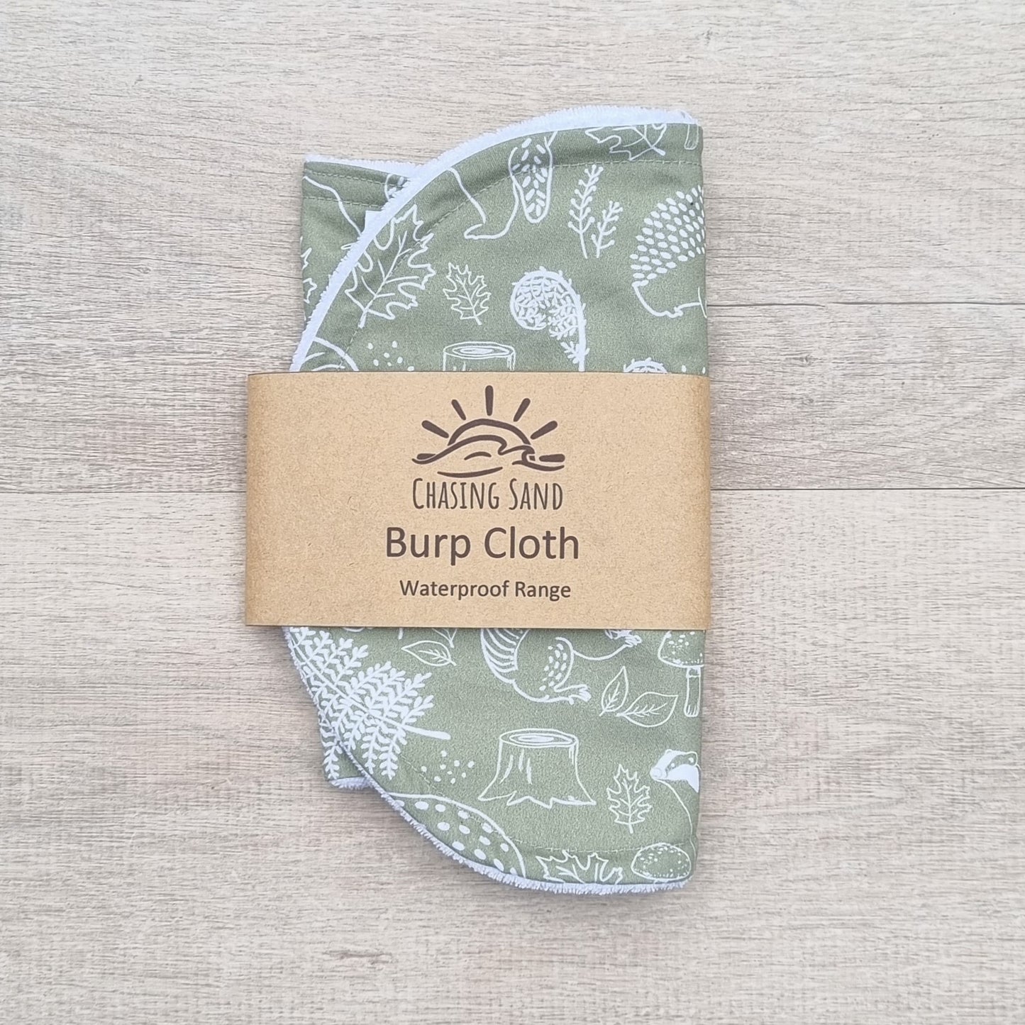 Burp Cloth - Into the Forest against wooden backdrop. White outlines of flora and fauna against sage green background.