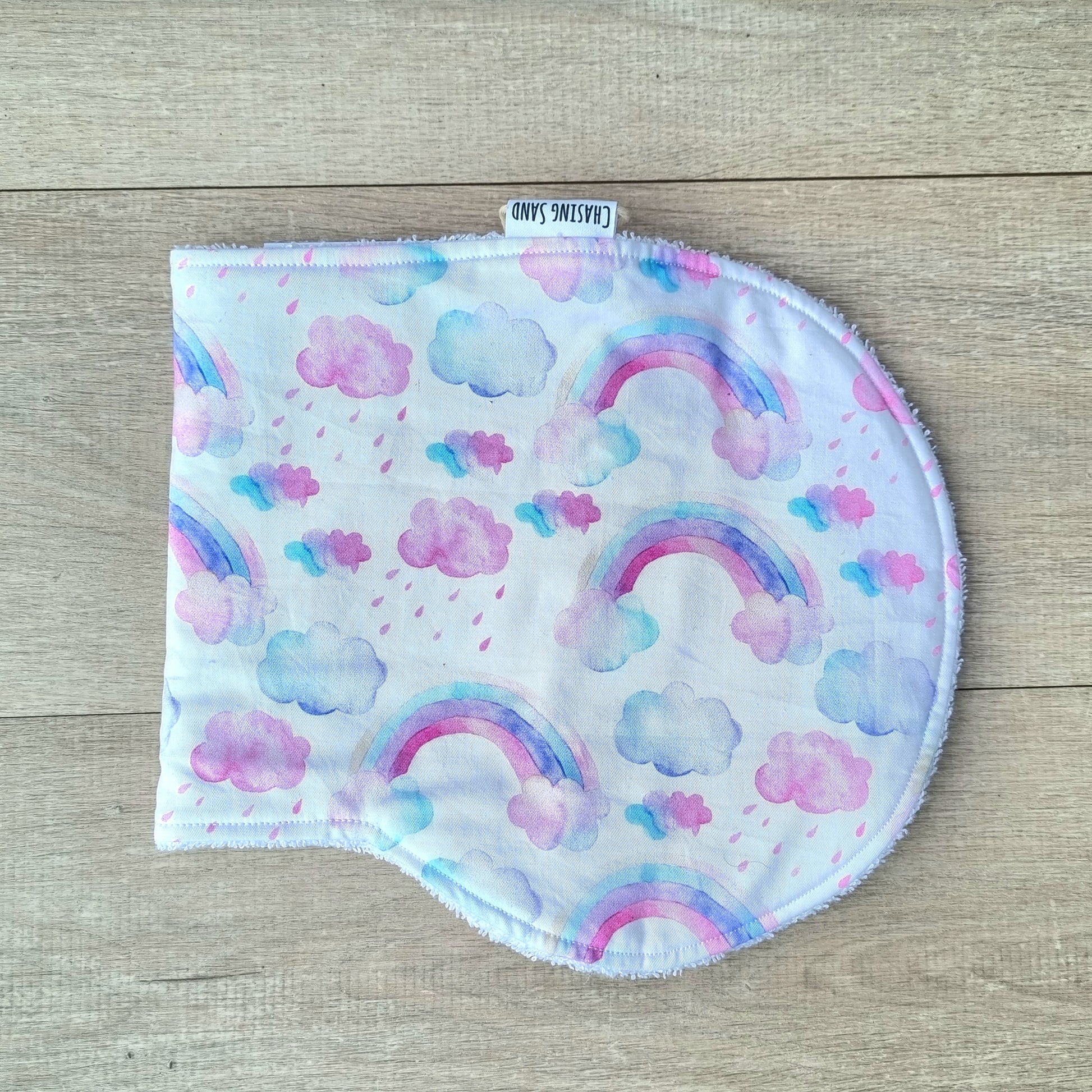 Burp Cloth - Pink Rainbows against wooden backdrop. Pink and purple watercolour rainbows and clouds against white background.