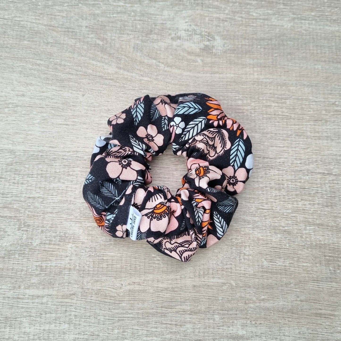 Scrunchie - Florence against wooden backdrop. Orange and pink flowers with leaves on black background.