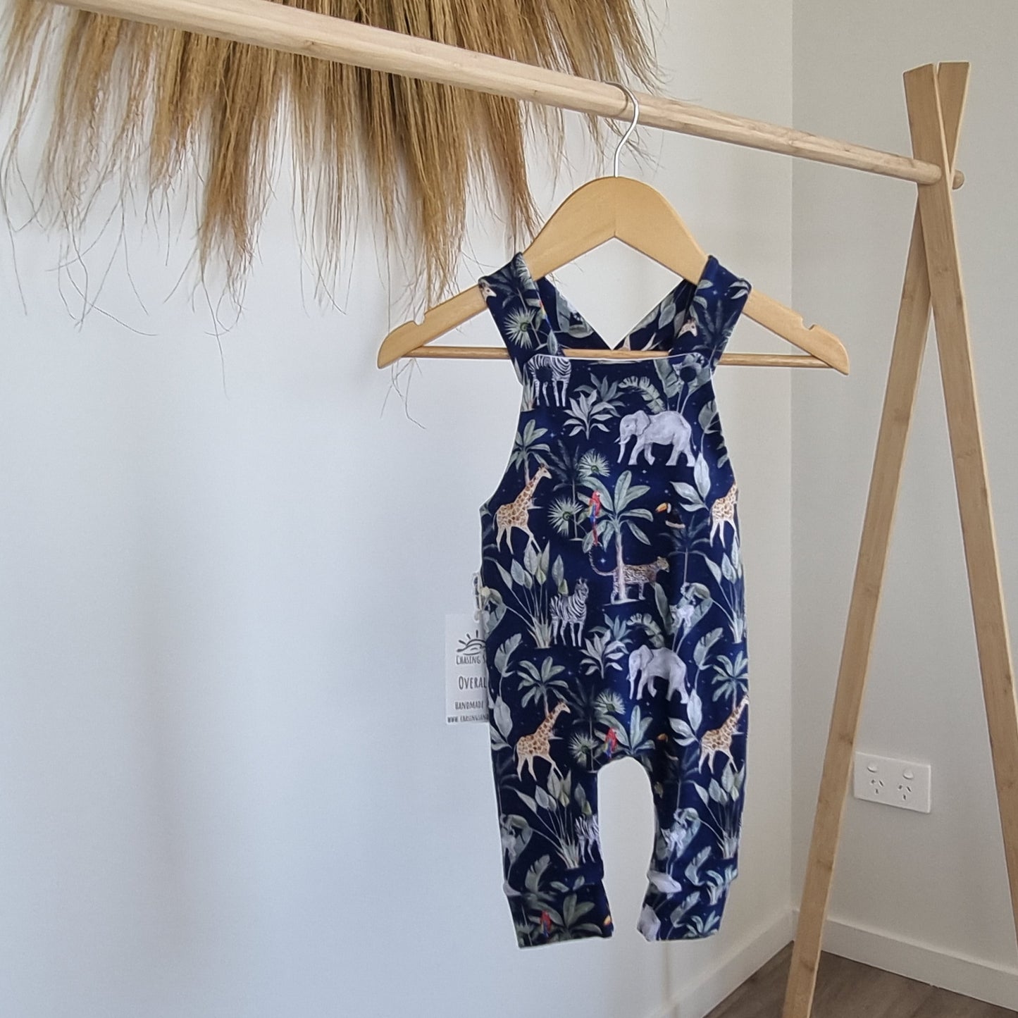 Long Overalls - Night Safari hanging on wooden rack. African animal and plant illustrations against navy blue background.