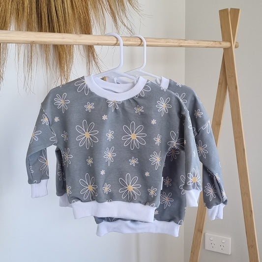 Jumper - Sweet Daisy hanging on a wooden rack. White and yellow daisies against soft grey background.