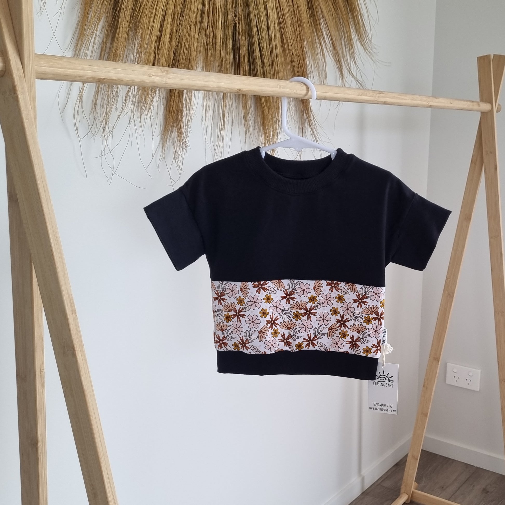 Tee - Sophie hanging on wooden rack. Black t-shirt with pattern around the middle: Brown, pink and orange flowers on white background