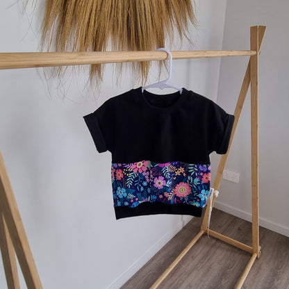Tee - Belle hanging on wooden rack. Black t-shirt with pattern around the middle: Pink, blue and purple flower design on navy background.