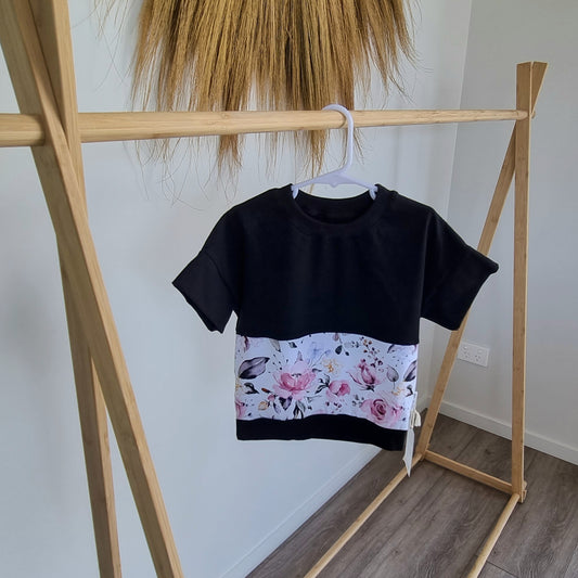 Tee - Rose Garden (Pink) hanging on wooden rack. Black t-shirt with pattern around the middle: .Pink watercolour rose design with green leaves on white background.