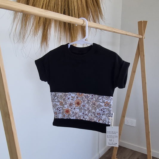 Tee - Avery hanging on wooden rack. Black t-shirt with pattern around the middle: Orange and yellow flower illustrations on white background.