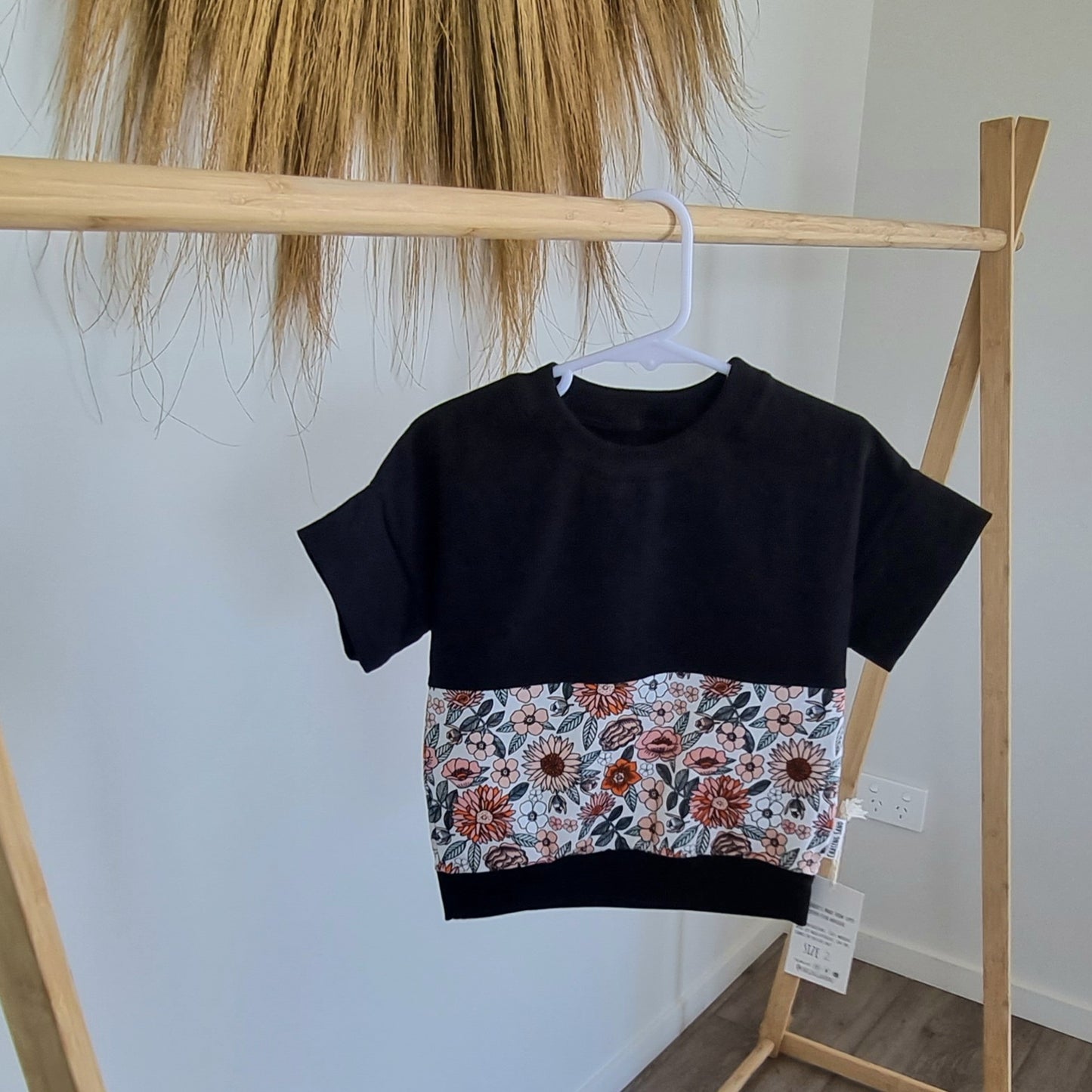 Tee - Florent hanging on wooden rack. Black t-shirt with pattern around the middle: Orange and peach flowers with green leaves on white background.