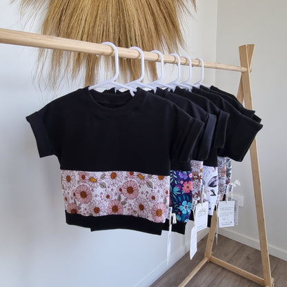 Tee - Harlow hanging on wooden rack. Black t-shirt with pattern around the middle: Pink and white flowers on white background.