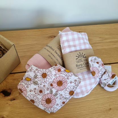 For Baby Gift Box - Welcome Baby Girl against wooden/white backdrop. Each Gift Box contains 1x Swaddle Blanket, 1x Burp Cloth, 1x Dribble Bib and 1x Teether.