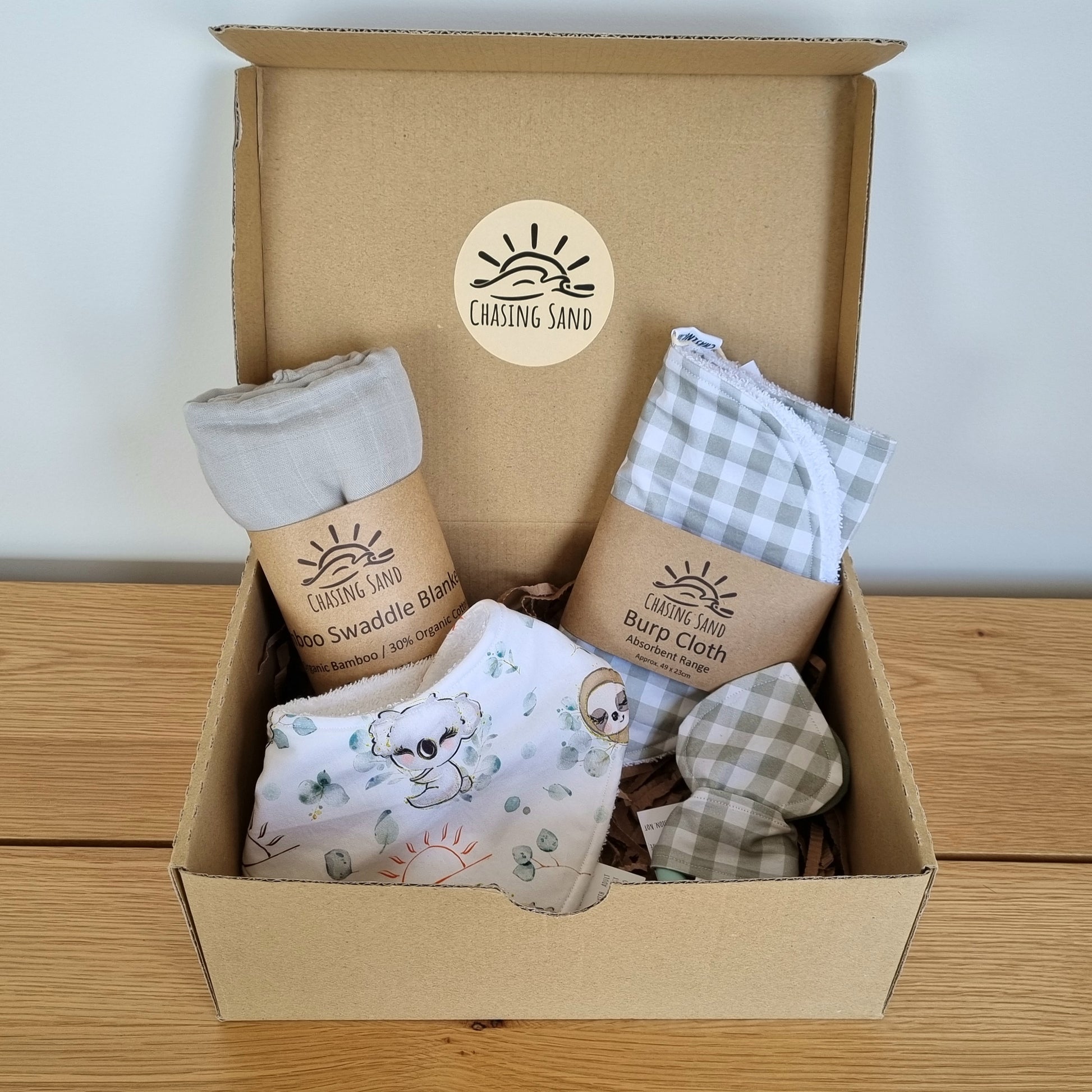 For Baby Gift Box - Neutral against wooden/white backdrop. Eco-friendly cardboard box containing 1x Swaddle Blanket, 1x Burp Cloth, 1x Dribble Bib and 1x Teether.