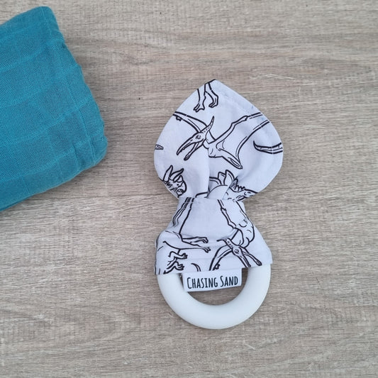 Teether -  B&W Dino against wooden backdrop. Black dinosaur design on white background, with white silicone teething ring.